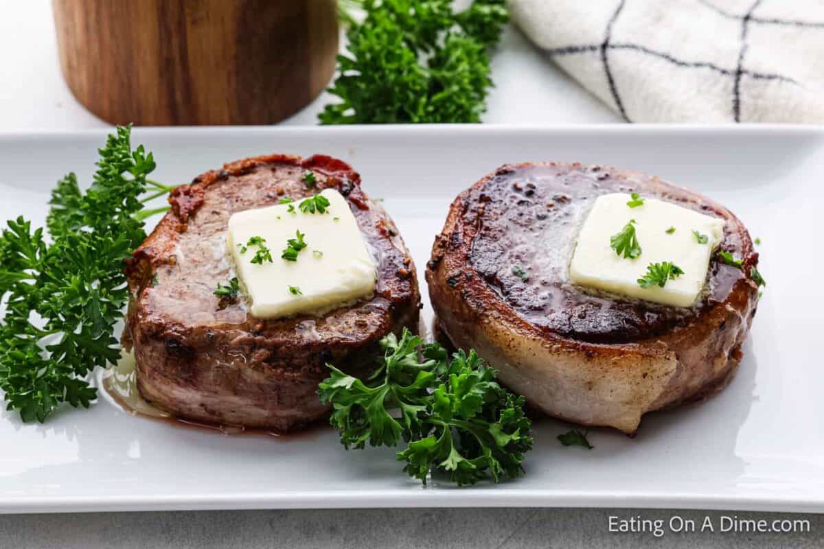 Two perfectly cooked bacon-wrapped filet mignon steaks, each topped with a pat of melting butter and garnished with fresh parsley, are served on a white rectangular plate. A sprig of parsley is placed alongside for decoration.