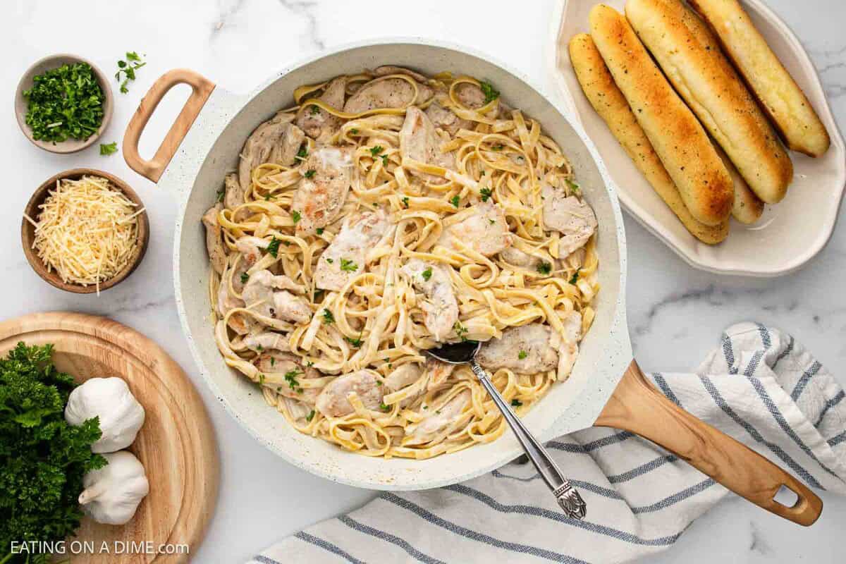 A skillet filled with a Creamy Chicken Alfredo Recipe sits on a striped towel, surrounded by breadsticks, shredded cheese, parsley, and garlic cloves on a marble countertop. A serving spoon rests in the skillet.