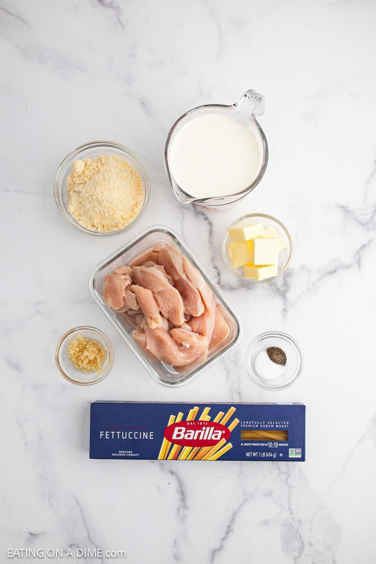 Top-down view of Creamy Chicken Alfredo Recipe ingredients on a white surface: raw chicken strips, Barilla fettuccine, grated parmesan, minced garlic, butter cubes, milk in a measuring cup, and a small bowl of black pepper.