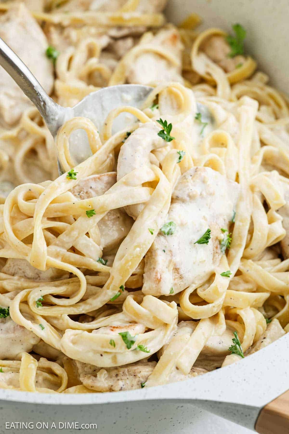 Close-up of a Creamy Chicken Alfredo Recipeโfettuccine pasta garnished with chopped parsley, being served with a metal spoon from a white pan.
