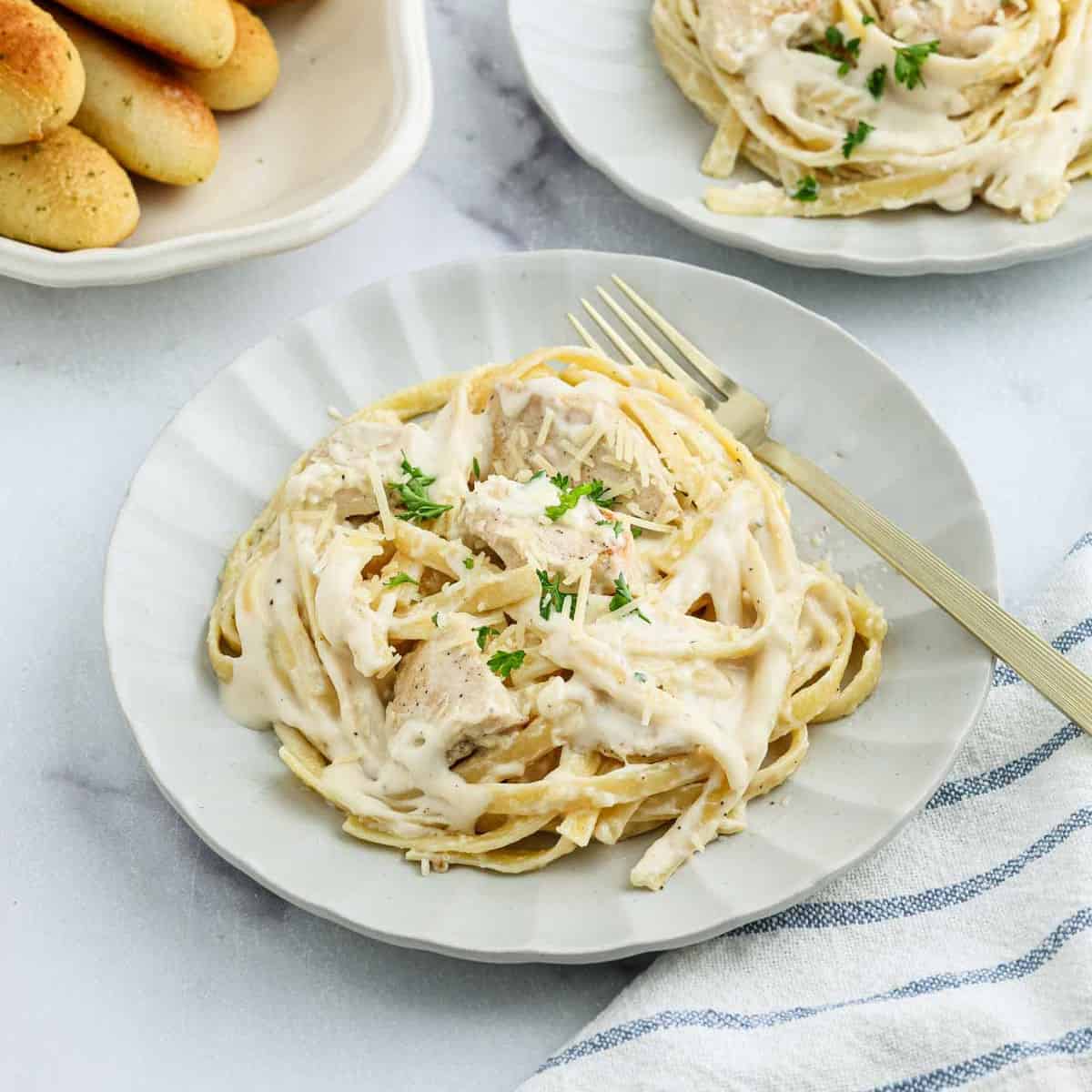 A plate of Creamy Chicken Alfredo Recipe fettuccine is topped with grated cheese and parsley. A gold fork rests on the plate, with breadsticks visible in the background.