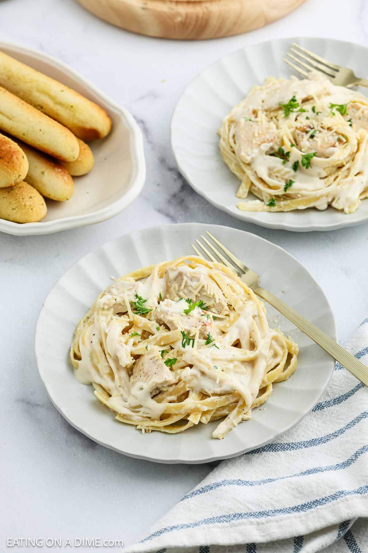 Two plates of Creamy Chicken Alfredo Recipe, topped with parsley and shredded chicken and served with forks, rest on a white table. A dish of breadsticks and a blue-striped napkin are set nearby for a cozy meal.