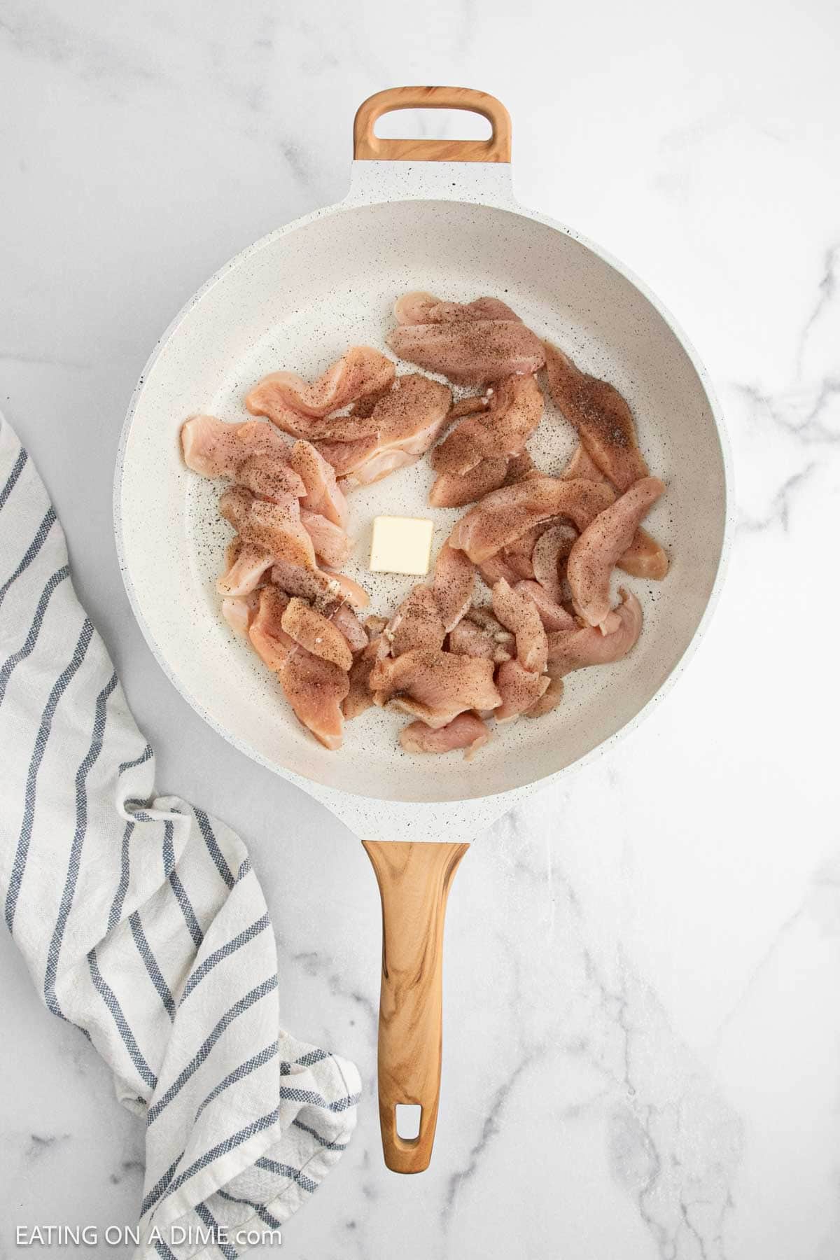 A white skillet with wooden handle holds raw, seasoned chicken strips and a pat of butter in the centerโperfect beginnings for a Creamy Chicken Alfredo Recipeโresting on a marble surface next to a striped kitchen towel.