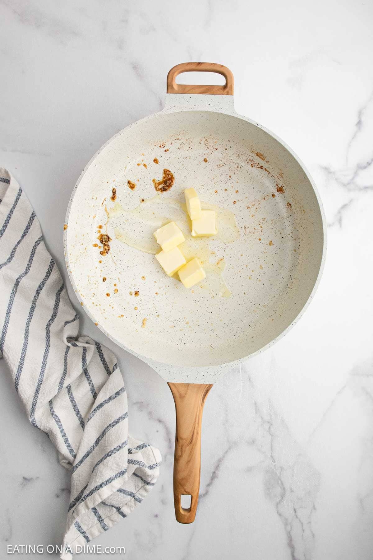 A white skillet with a wooden handle contains several cubes of butter beginning to meltโperfect for starting a Creamy Chicken Alfredo Recipe. A striped kitchen towel lies beside the skillet on a white marble surface.