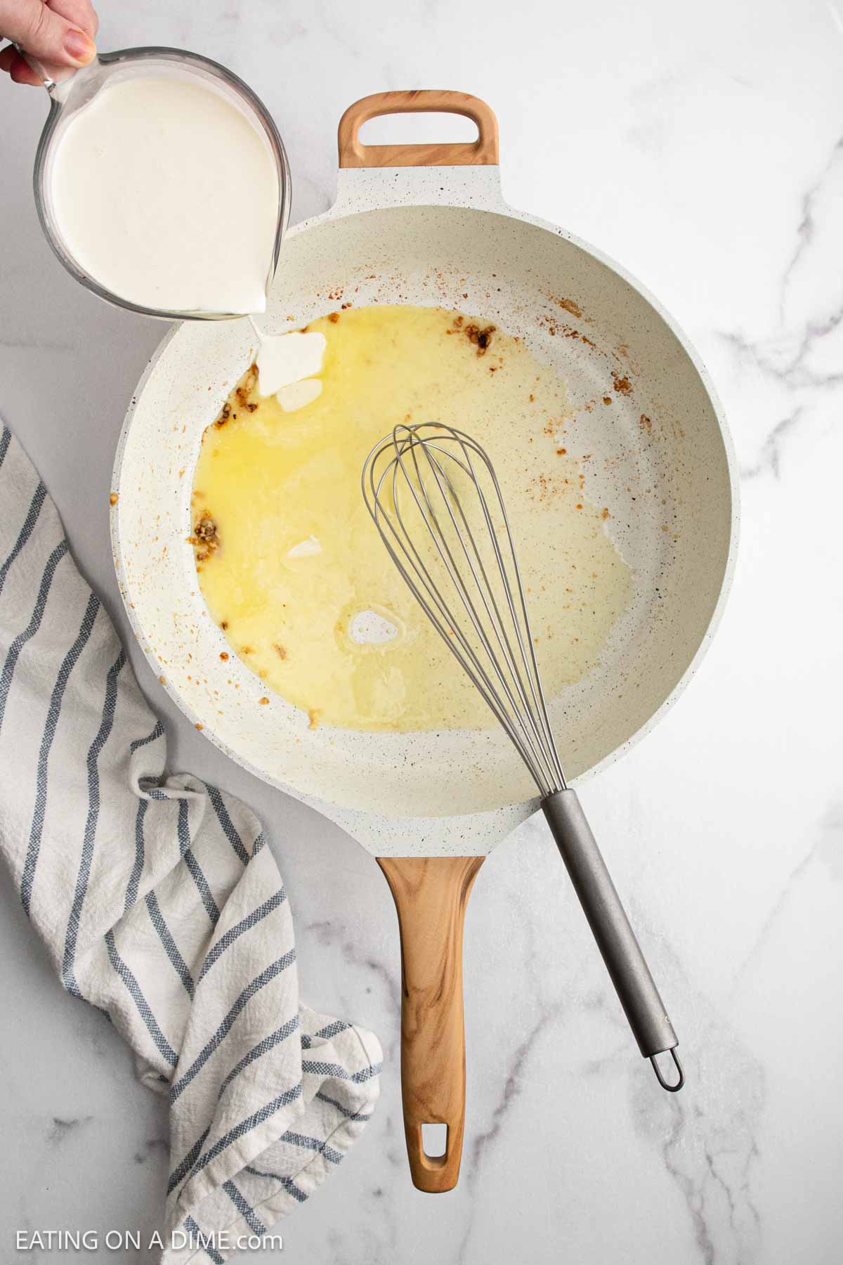 A hand pours cream from a measuring cup into a white skillet with melted butter and a whisk on a marble countertopโa classic step in making Creamy Chicken Alfredo Recipe. A striped kitchen towel is nearby.