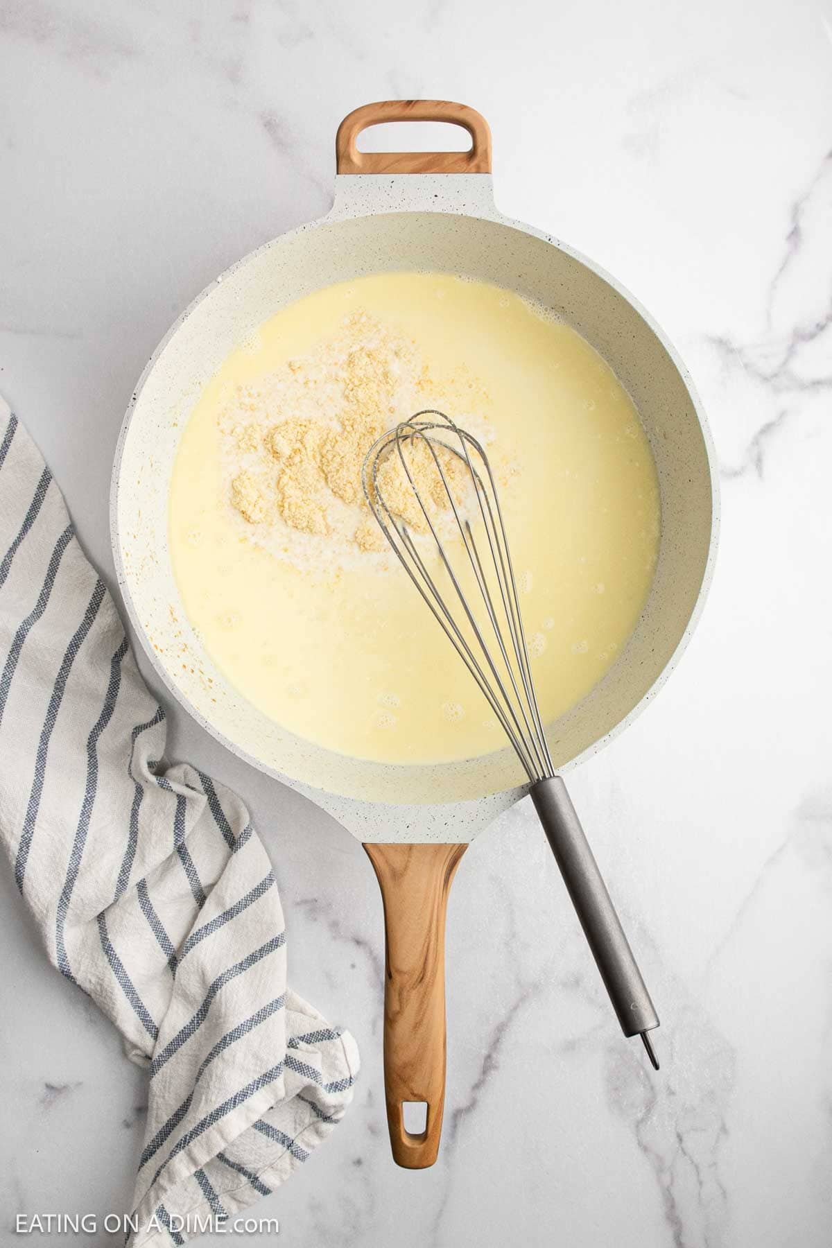 A light-colored skillet with a wooden handle contains a creamy liquid and some flour, being mixed with a metal whiskโperfect for starting a Creamy Chicken Alfredo Recipe. A striped dish towel lies beside the skillet on a marble surface.