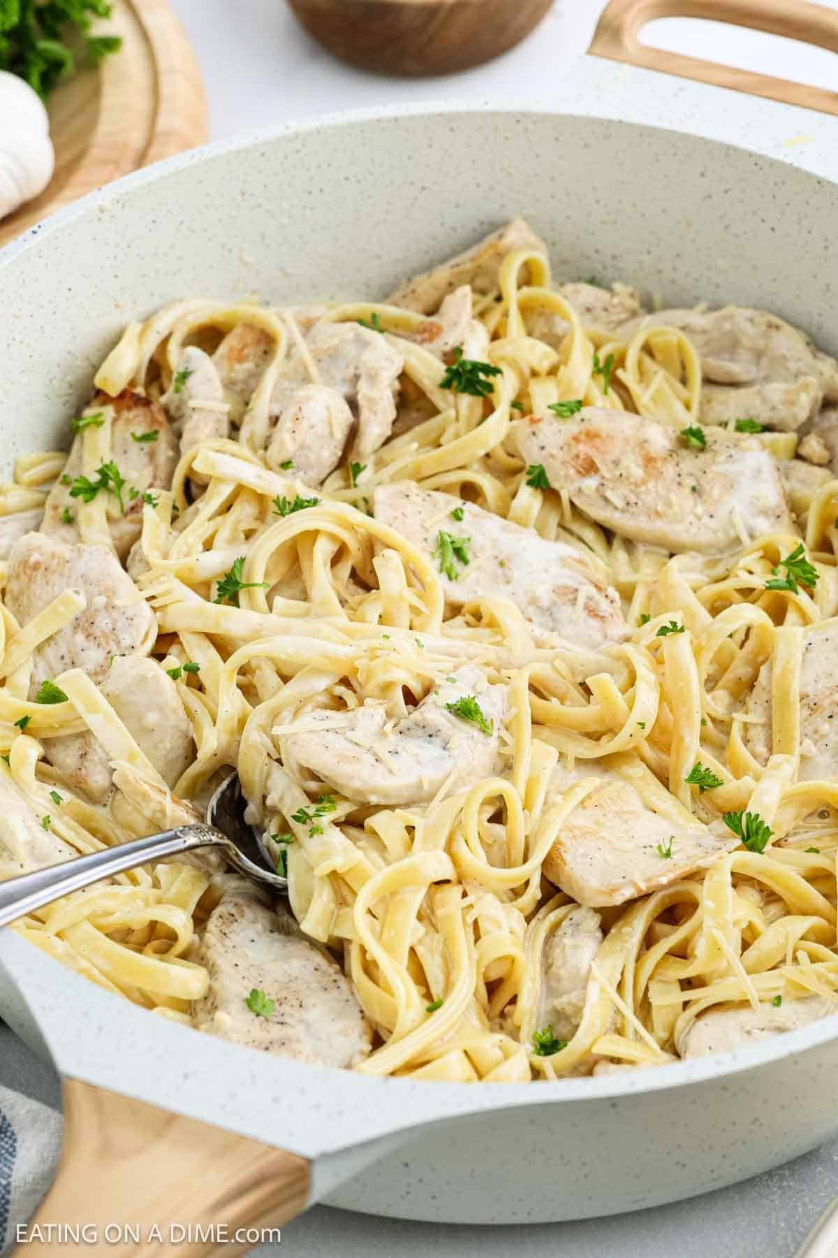 A large white skillet filled with creamy fettuccine pasta and sliced chicken, garnished with chopped parsleyโa delicious Creamy Chicken Alfredo Recipe. A silver serving fork is resting in the pan.