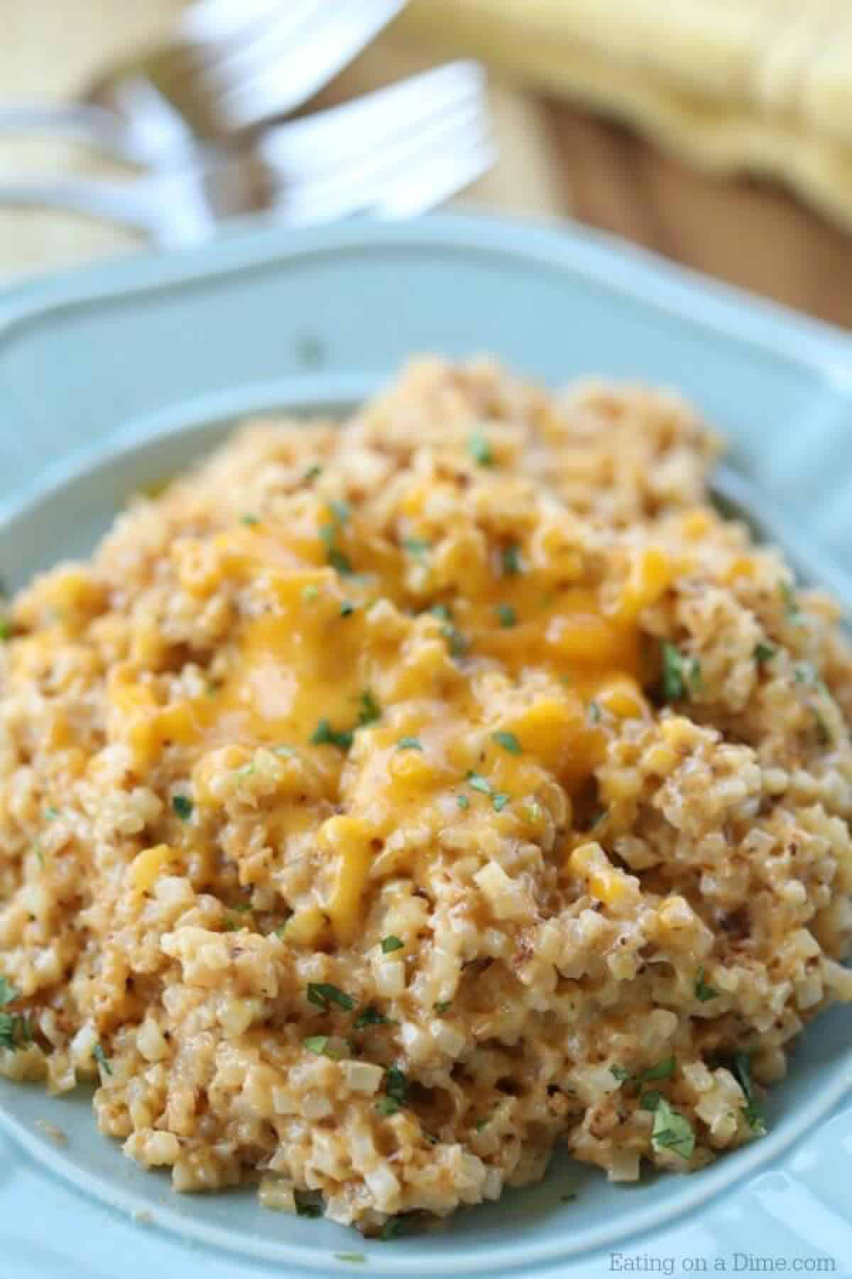 Looking for an easy keto side dish? You're going to love Easy Cheesy Cauliflower Rice. With just a few ingredients you can have Keto Cheesy Cauliflower. Close up image of cheesy cauliflower rice in a blue bowl topped with cheese.