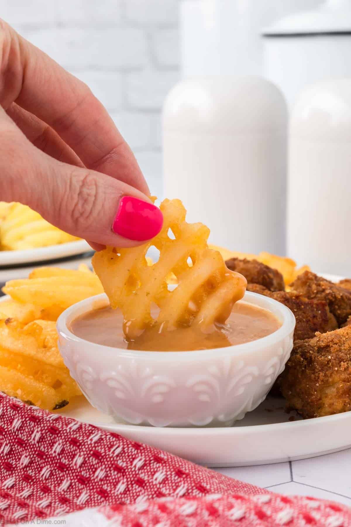 A hand dipping waffle french fries into a bowl of Honey Roasted BBQ Sauce on a plate of waffle fries and fried chicken nuggets