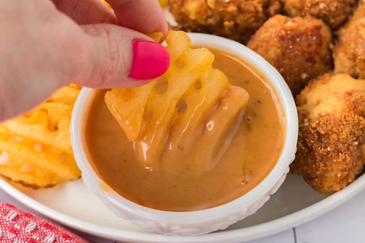 A hand dipping waffle fries into a bowl of Honey BBQ Sauce on a plate of fried chicken nuggets