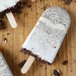 A cookies and cream popsicle with visible cookie pieces sits on a wooden surface, surrounded by cookie crumbs, showcasing the classic appeal of cookies and cream popsicles.