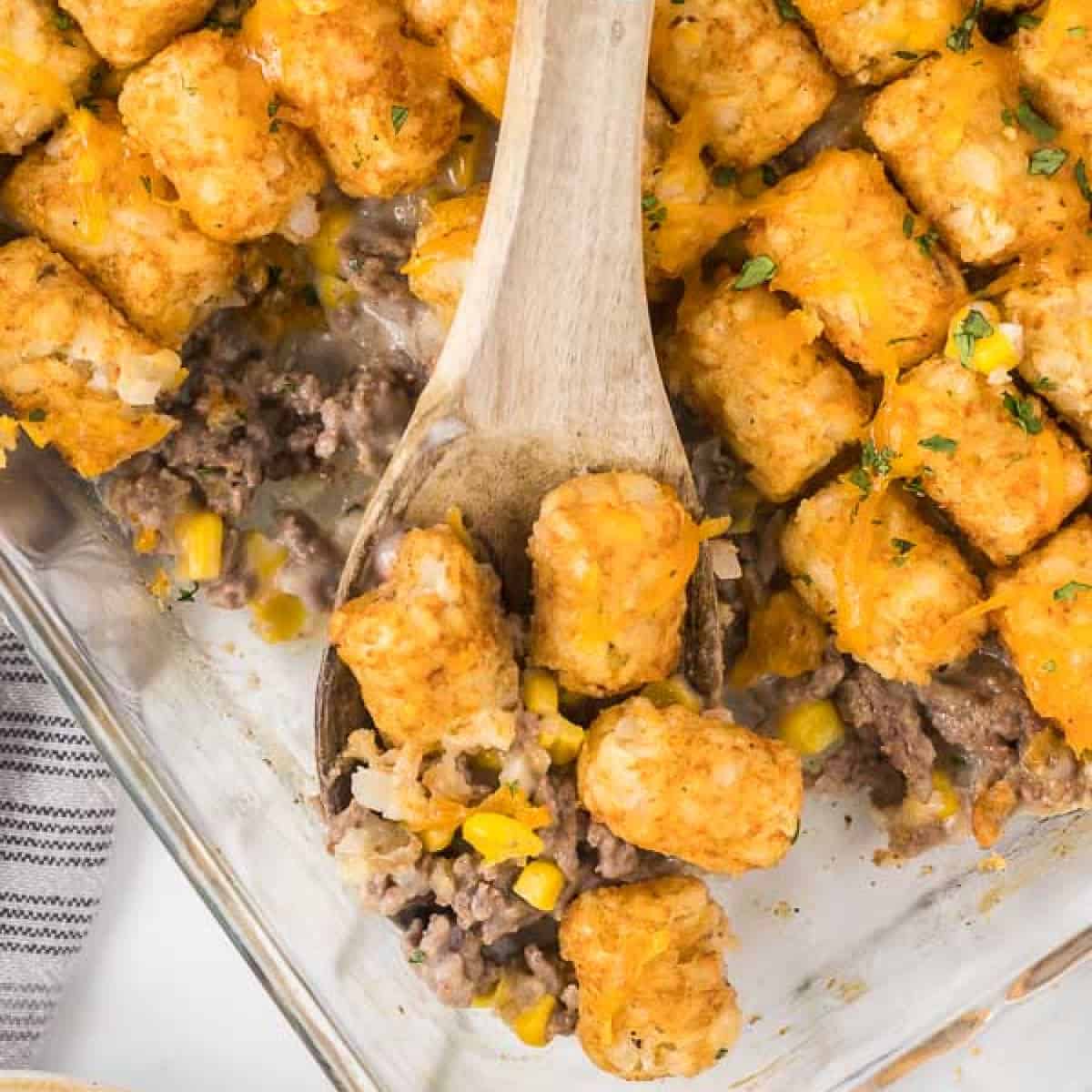 A close-up of Cowboy Casserole features a dish brimming with tater tots, ground beef, corn, and melted cheese. A wooden spoon scoops out a hearty portion, while chopped herbs add the perfect garnish to this rustic delight.