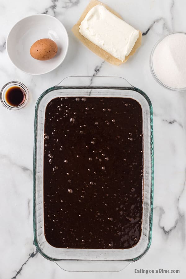Brownie Batter in a baking dish with a bowl with a egg, vanilla extract, cream cheese and sugar