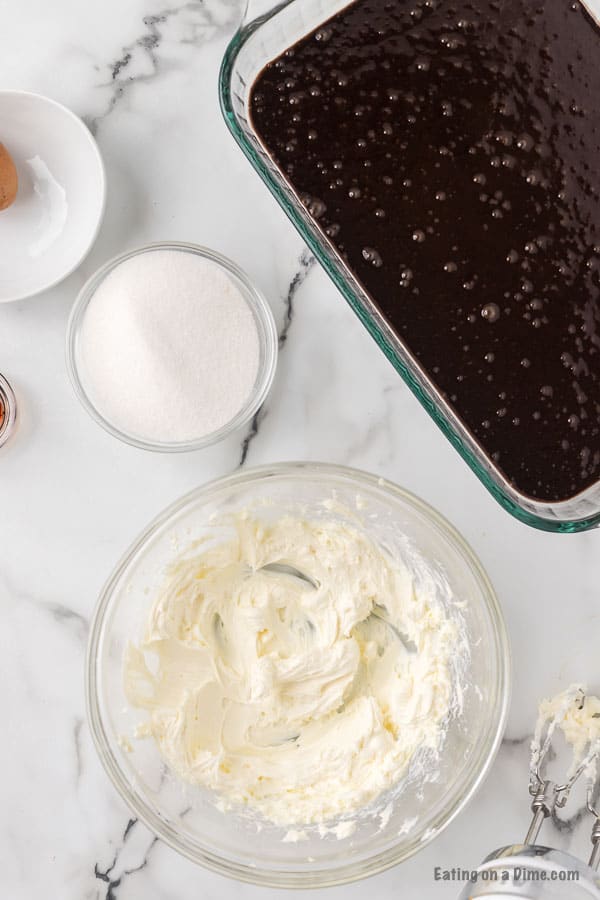 Bowl with creamed cream cheese and a bowl of sugar and the baking dish with brownie batter
