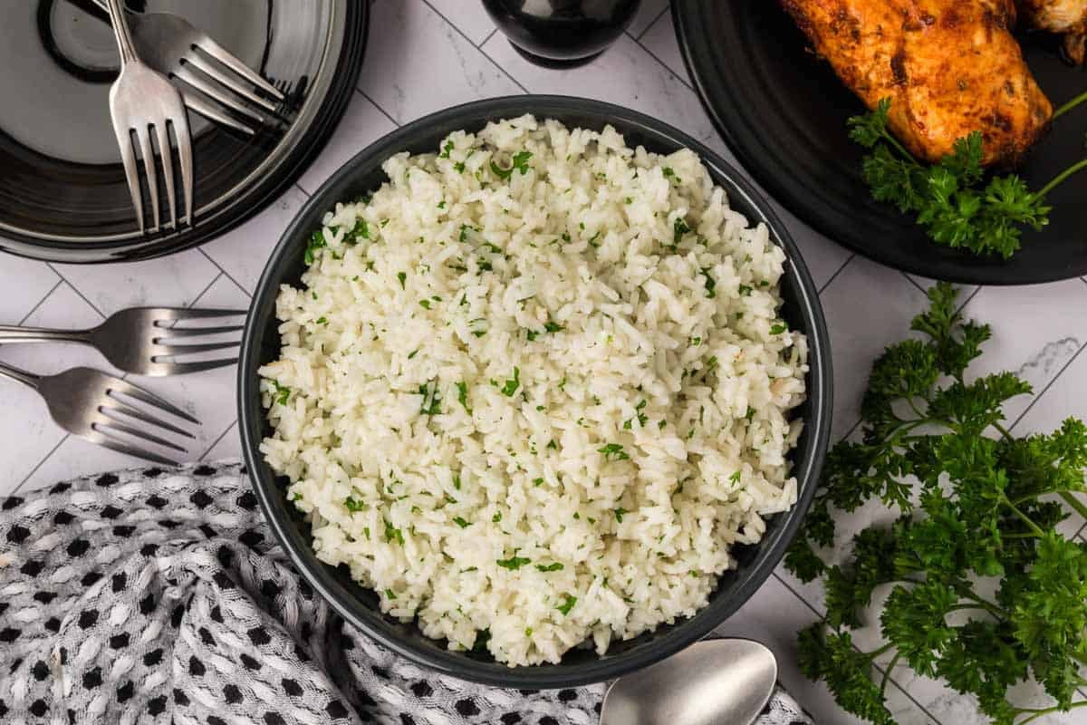 A black bowl filled with cooked white rice garnished with chopped parsley, placed on a table with plates, forks, a spoon, a patterned cloth, and fresh parsley nearby—perfect for learning how to season white rice.
