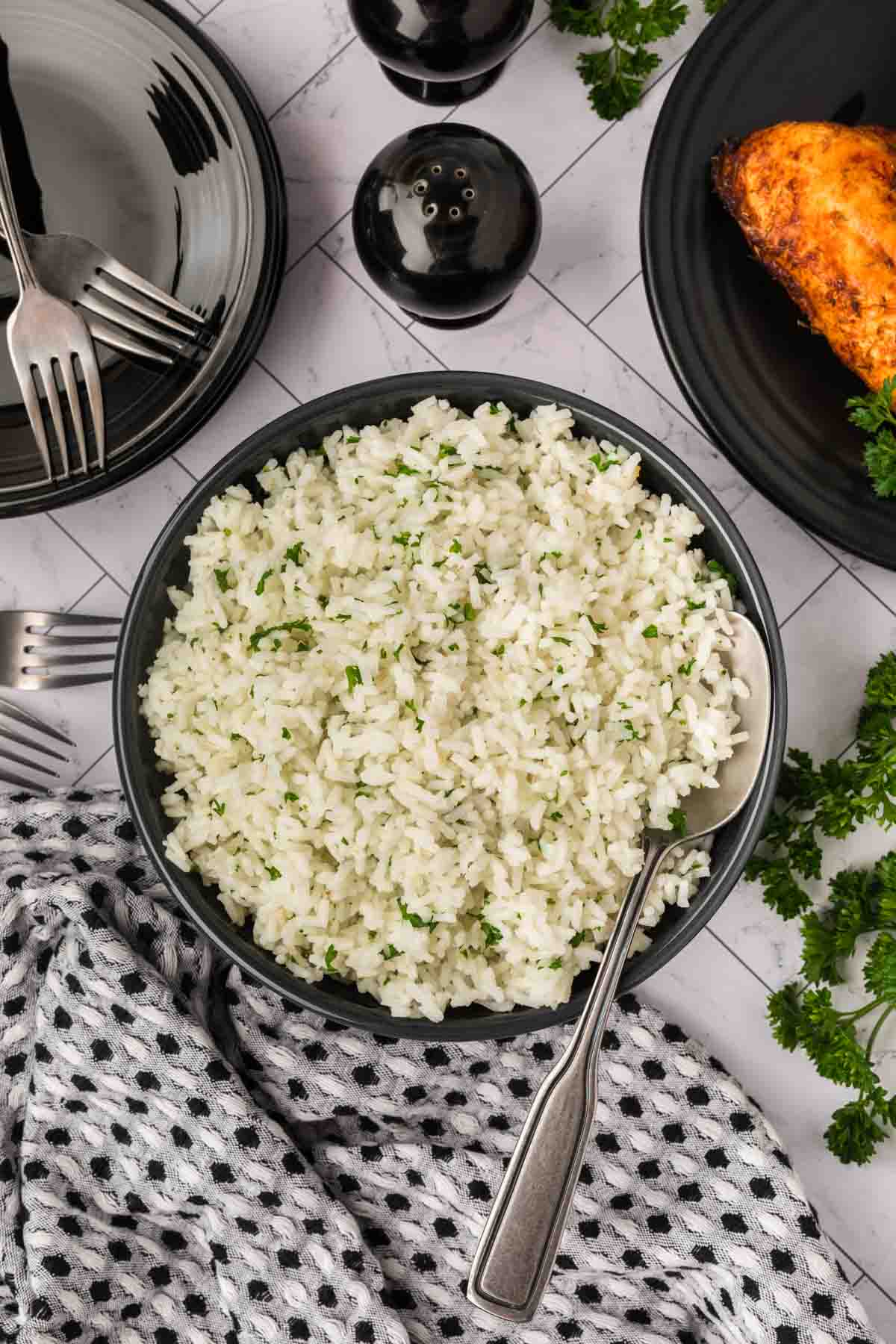 A bowl of white rice garnished with chopped herbs, showing How to Season White Rice, with a spoon inside. Plates, forks, a napkin, salt and pepper shakers, and roasted chicken are arranged nearby on a white surface.