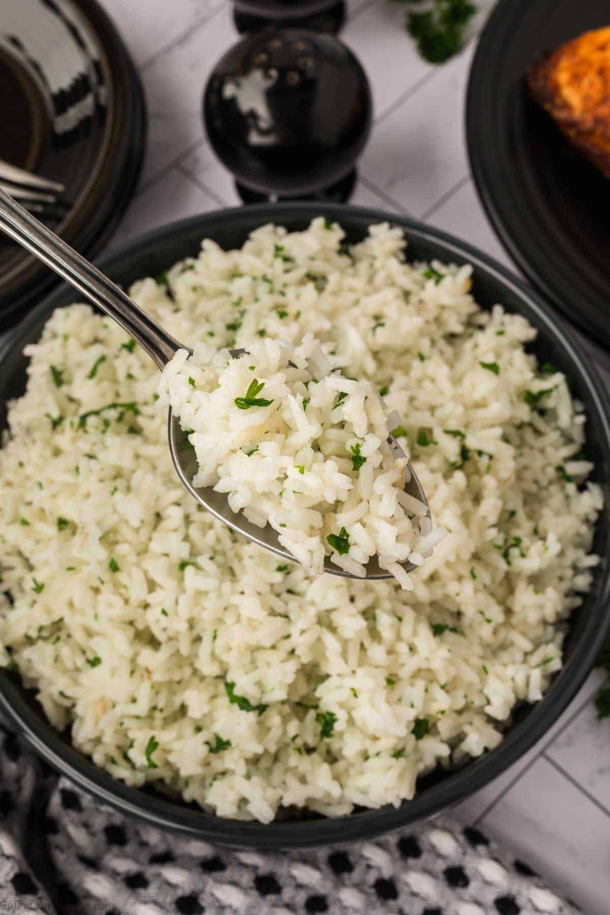 A bowl of fluffy white rice garnished with chopped parsley, with a spoon lifting a portion above the bowl. Wondering how to season white rice? A dark plate and salt and pepper shakers are visible in the background.