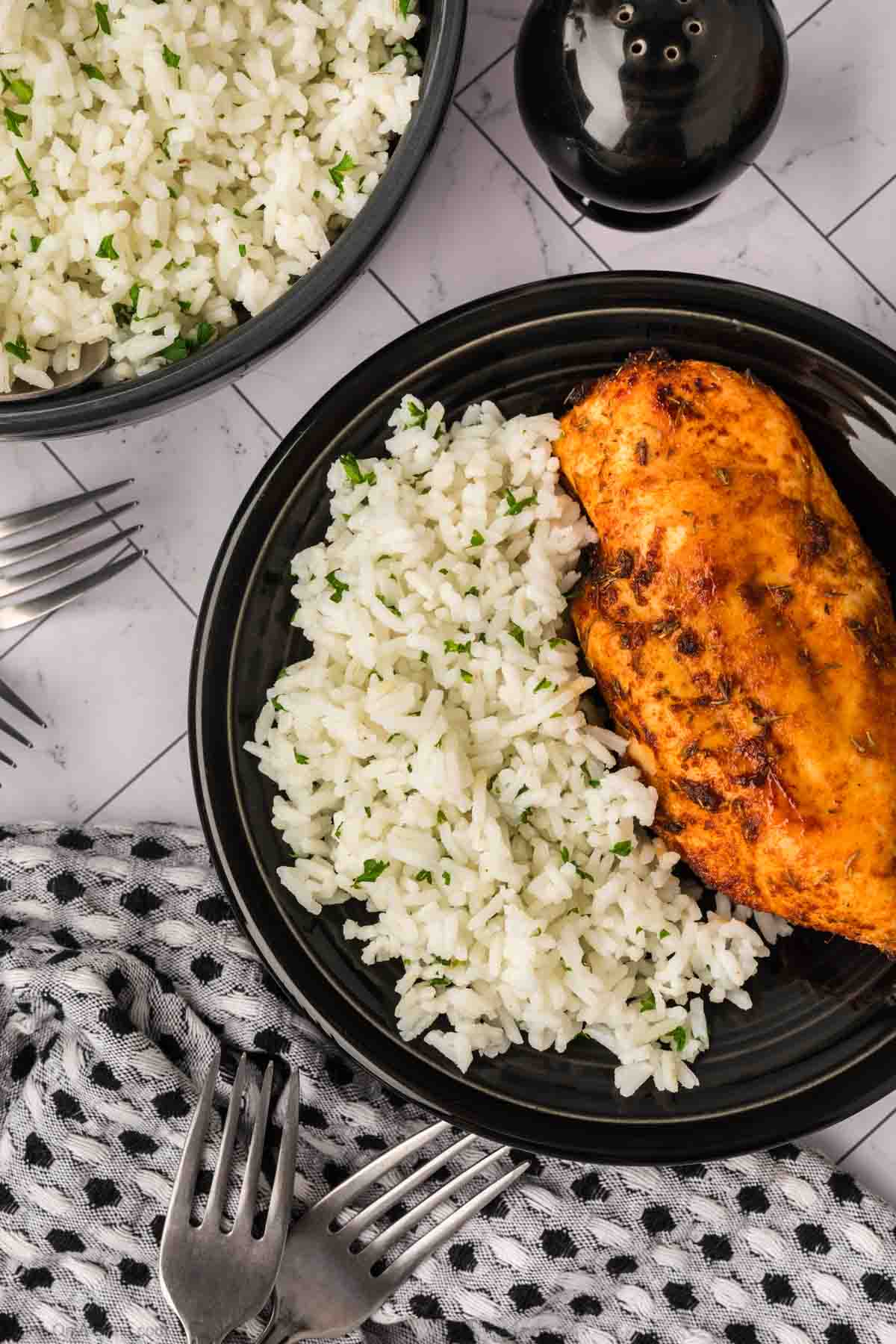 A black plate with a serving of white rice garnished with herbs—showcasing How to Season White Rice—and a seasoned, cooked chicken breast, next to a bowl of rice, forks, and a black-and-white napkin on a tiled surface.