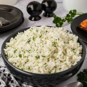 A black bowl filled with fluffy white rice garnished with chopped herbs sits on a table with black plates, cutlery, a napkin, salt and pepper shakers—ideal for those curious about how to season white rice perfectly.