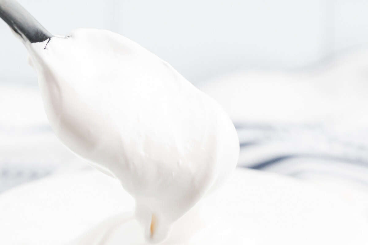 A close-up of a spoon holding a dollop of thick, white whipped cream or meringue, perfect for your next Marshmallow Creme Recipe, with a soft-focus background.