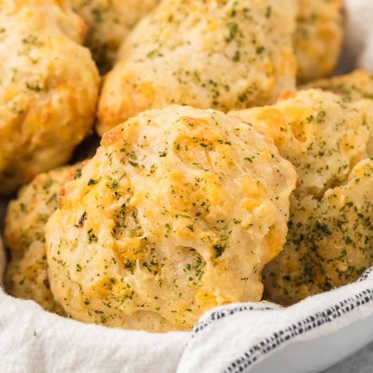 Close-up of golden-brown, fluffy Cheddar Bay Biscuits sprinkled with herbs, piled on a white cloth. The textured surface of these Red Lobster favorites is arranged in a bowl, highlighting their savory appearance.