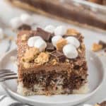 A close-up of a S'mores Cake dessert bar topped with mini marshmallows, chocolate pieces, and graham cracker crumbs on a white plate, with a fork beside it and more dessert bars in the background.