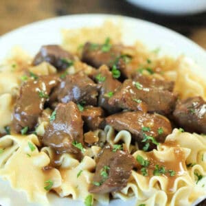 A plate of wide egg noodles topped with tender pieces of beef in a rich brown gravy, garnished with chopped parsley, presented on a white plate.