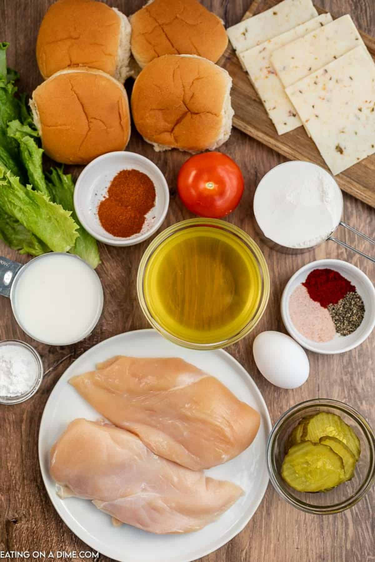 Ingredients for a sandwich reminiscent of a Chick-Fil-A Spicy Deluxe Sandwich are laid out on a wooden surface, featuring chicken breasts, sandwich buns, lettuce, tomato slices, cheese, pickles, flour, seasonings, an egg mixture with milk, and a bowl of oil ready for frying.