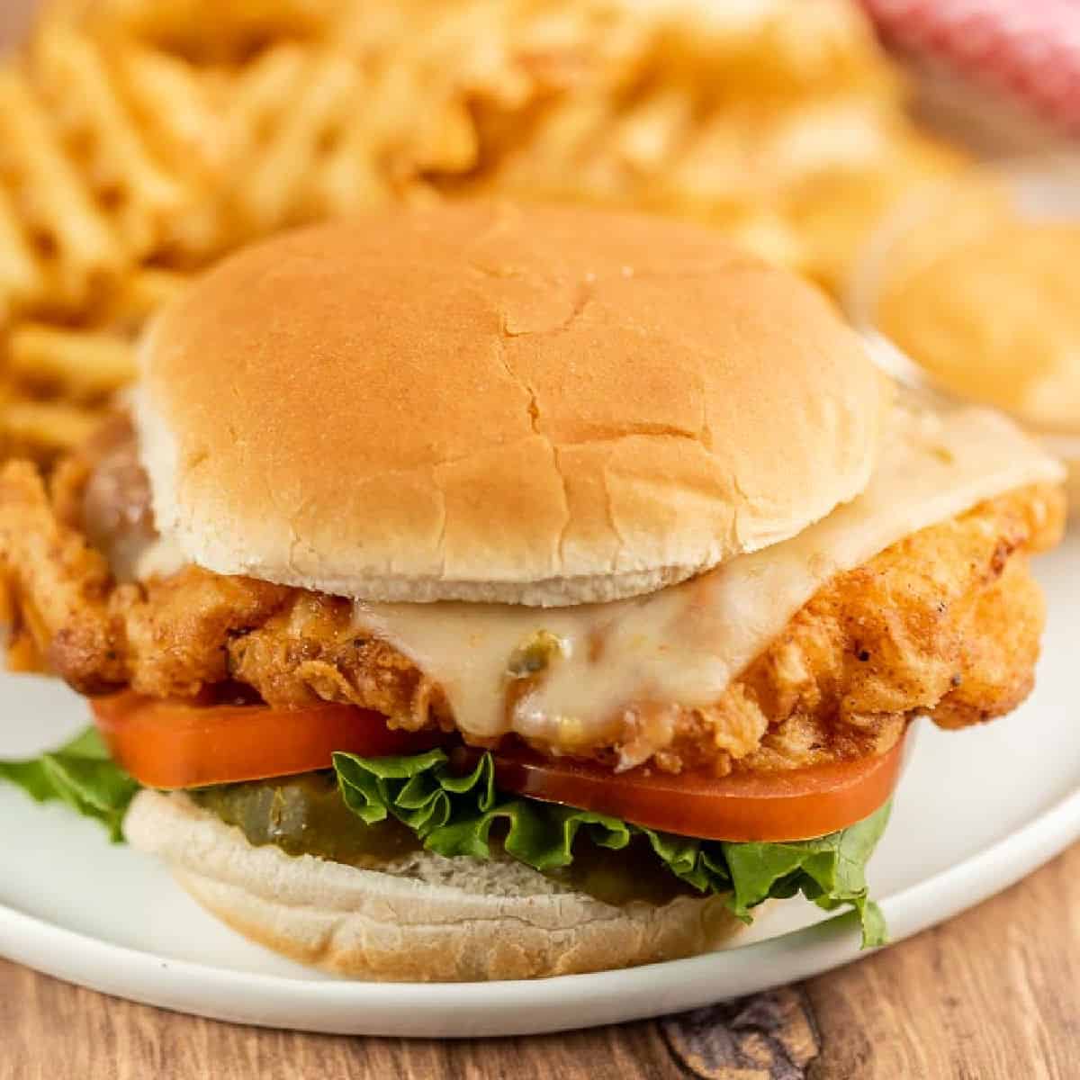 A close-up of a Chick-Fil-A Spicy Deluxe Sandwich on a white plate. The sandwich features a crispy fried chicken patty with lettuce, tomato slices, pickles, and melted cheese inside a soft bun. Waffle fries and dipping sauce are artfully blurred in the background.
