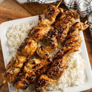 Teriyaki chicken on a stick, glazed and sprinkled with sesame seeds, served over a bed of white rice on a square white plate.