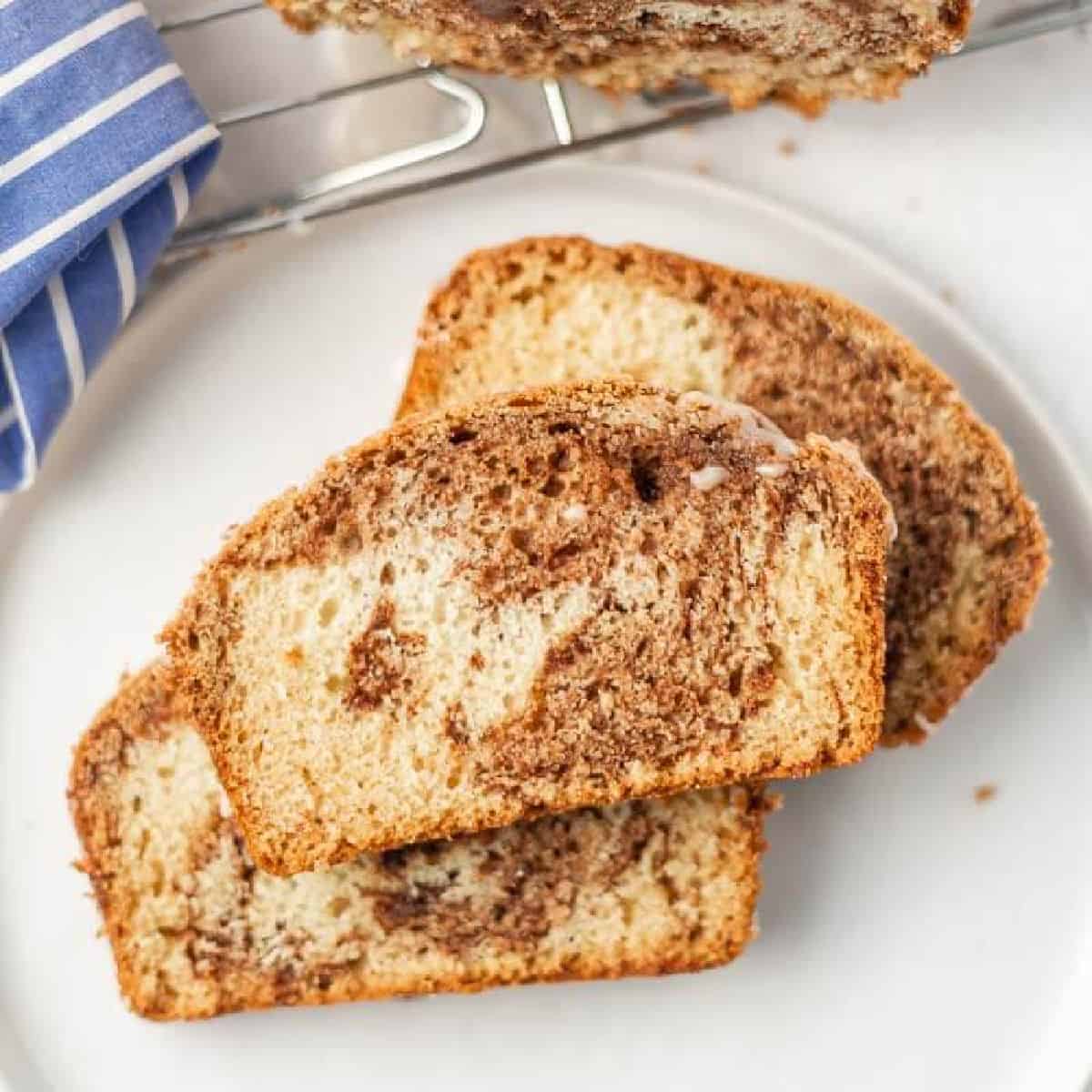 Three slices of marbled cinnamon swirl bread, made from a classic cinnamon swirl bread recipe, are arranged on a white plate. The bread features a beautiful pattern of light and dark brown. A striped blue cloth and cooling rack appear in the background.