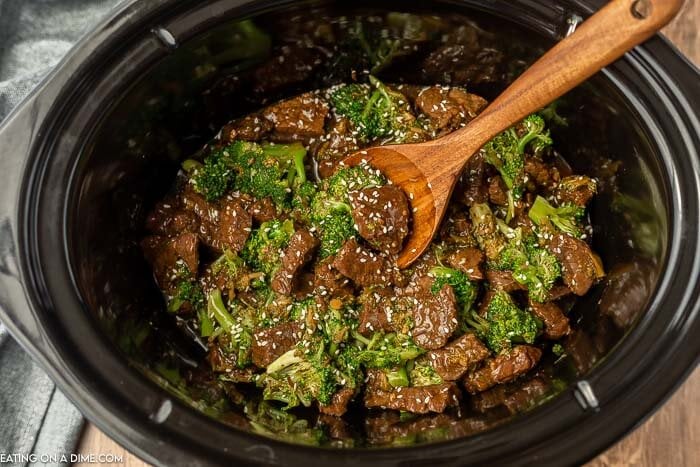 Close up image of beef and broccoli in a crock pot with a wooden spoon. 