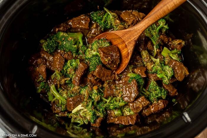 Close up image of beef and broccoli in a crock pot with a wooden spoon. 