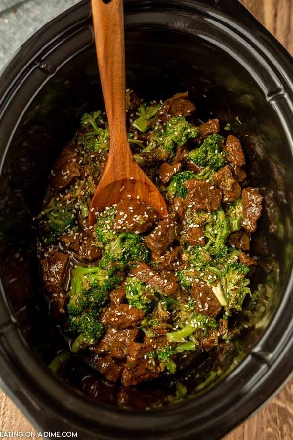 Close up image of beef and broccoli in a crock pot with a wooden spoon. 