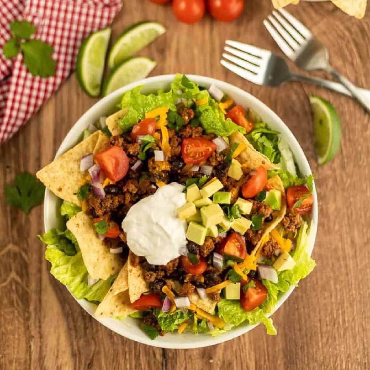 A bowl of Crockpot Taco Salad Recipe with ground beef, lettuce, cherry tomatoes, black beans, cheese, avocado, red onion, tortilla chips, and a dollop of sour cream. Two forks, lime wedges, and a red napkin sit nearby on a wooden table.