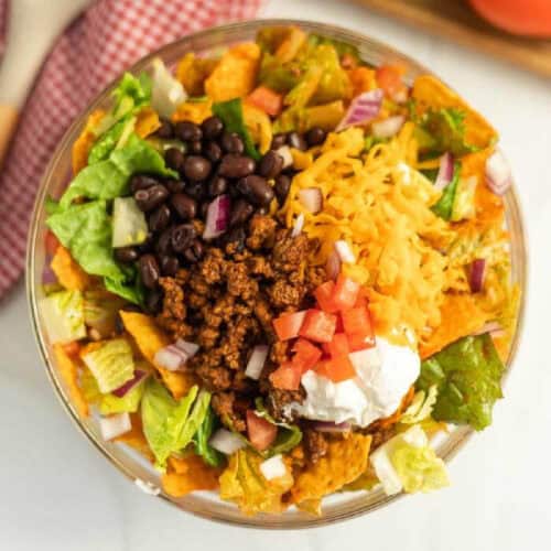 A vibrant Doritos Taco Salad in a clear bowl features seasoned ground beef, black beans, shredded cheddar cheese, chopped tomatoes, diced onions, lettuce, and a dollop of sour cream on top.