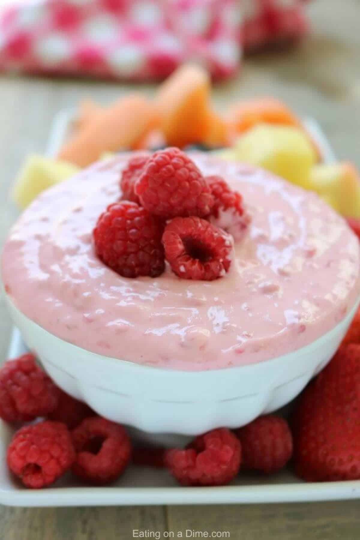 Learn how to make this easy and delicious Raspberry fruit dip recipe. You only need 4 ingredients to make Raspberry cream cheese dip recipe. It goes well with any fruit and is sure to be a hit! Serve Raspberry cream cheese fruit dip recipe at parties, BBQ's and more. It's the perfect snack anytime! A bowl of raspberry fruit dip topped with fresh raspberries with more fresh fruit behind it for serving.