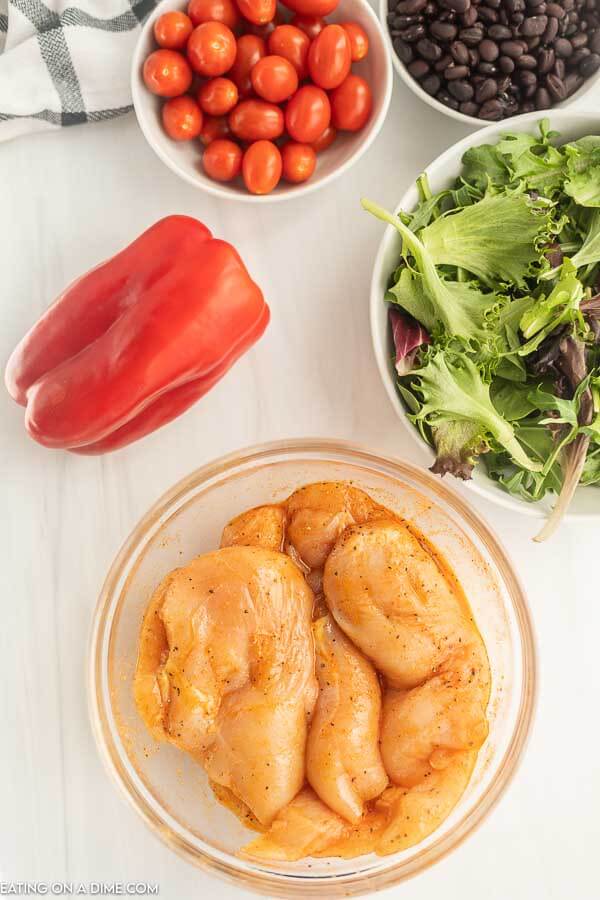 Close up image of chicken marinating in a clear bowl with a red bell pepper and a bowl of greens. 