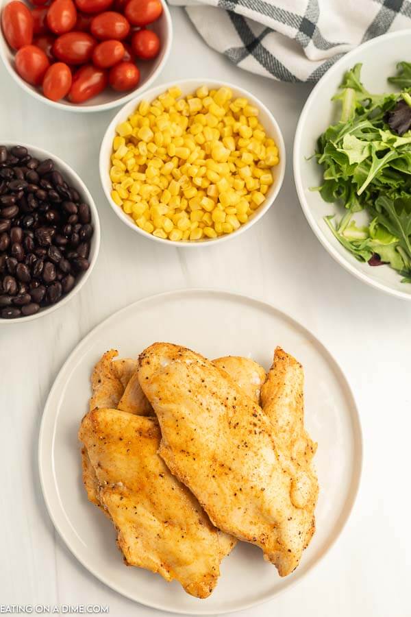 Close up image of cook chicken on a plate, a bowl of corn, black beans, grape tomatoes, and salad