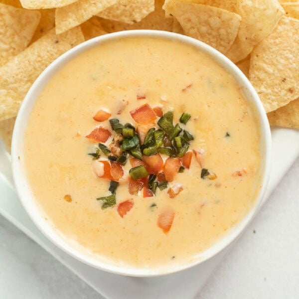 A bowl of creamy Copycat Qdoba Queso Recipe garnished with chopped tomatoes and green herbs, surrounded by crispy tortilla chips on a white plate.