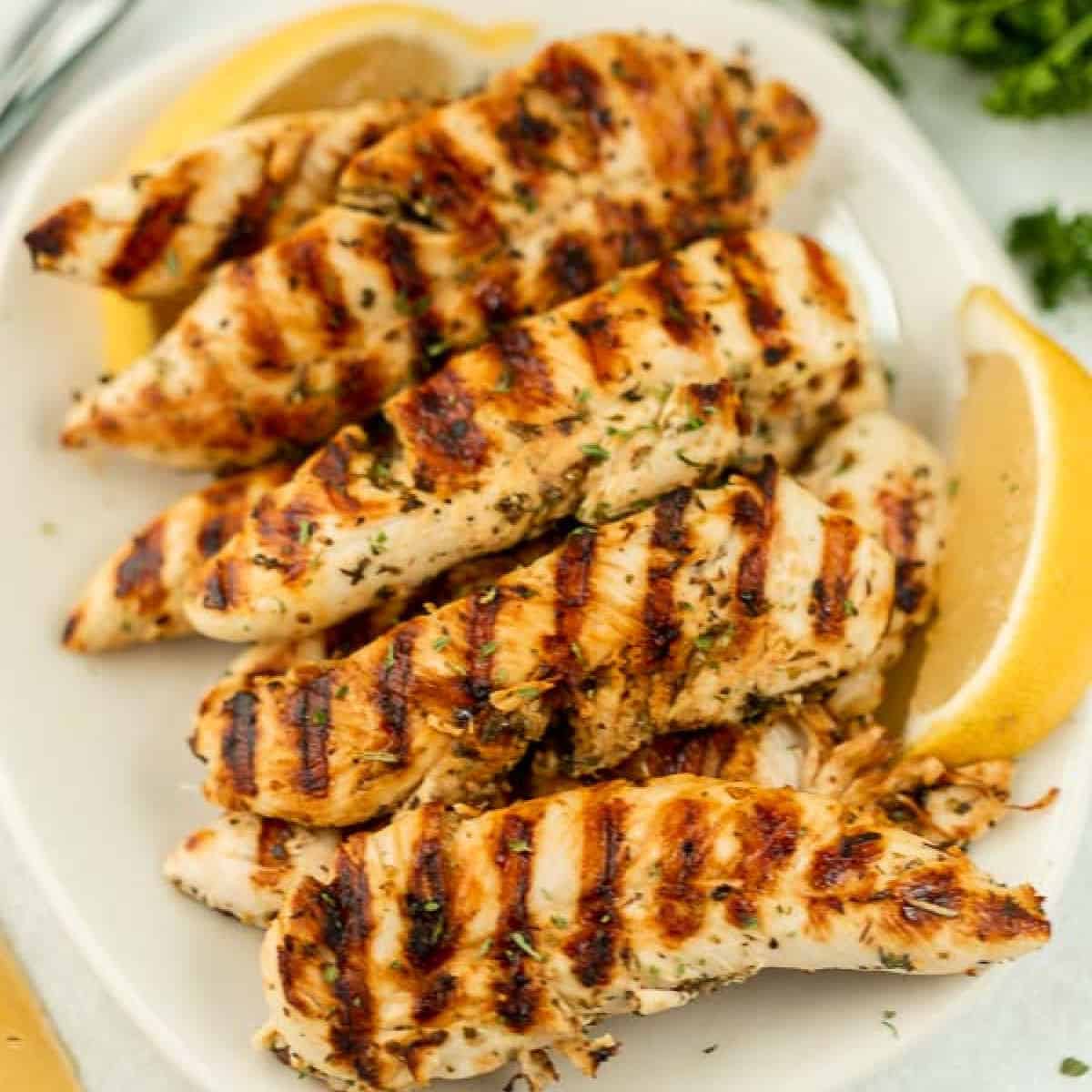 Grilled chicken tenders garnished with herbs on a white plate, accompanied by lemon wedges.