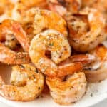 Close-up of garlic parmesan shrimp seasoned with herbs and grated Parmesan cheese, arranged on a white plate. The shrimp appear golden and slightly crispy, with visible seasoning and a garnish of parsley.