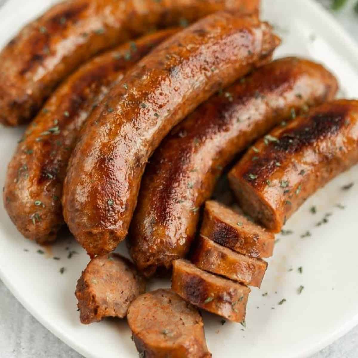 A plate with several cooked sausages, some whole and others sliced, garnished with herbs. The enticingly browned and glistening Italian sausages are expertly arranged neatly on a white plate, showcasing how to cook Italian sausage to perfection.