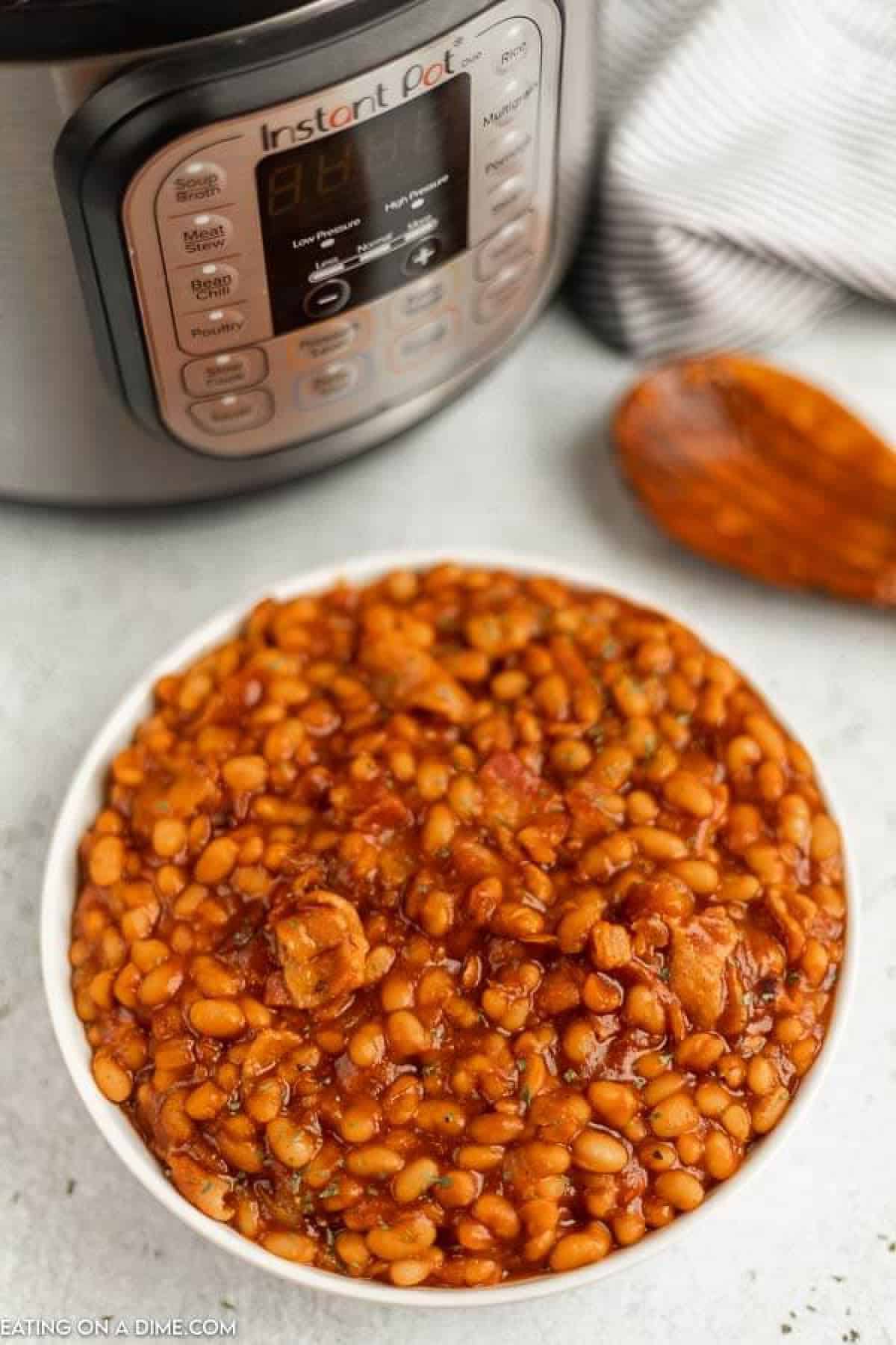 Close up image of baked beans in a white bowl with an instant pot and a wooden spoon.