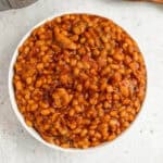 A bowl filled with Instant Pot baked beans cooked in a rich, reddish-brown sauce. The dish appears to have small pieces of meat and herbs mixed in. The background is a light, speckled surface.