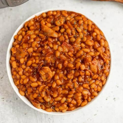 A bowl filled with Instant Pot baked beans cooked in a rich, reddish-brown sauce. The dish appears to have small pieces of meat and herbs mixed in. The background is a light, speckled surface.