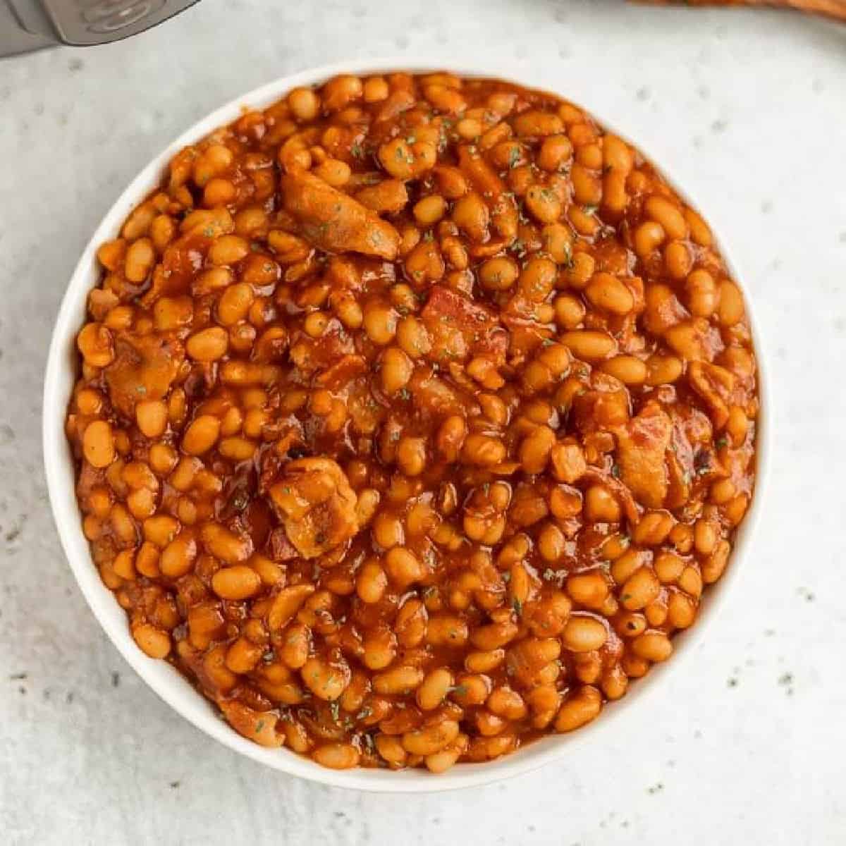 A bowl filled with Instant Pot baked beans cooked in a rich, reddish-brown sauce. The dish appears to have small pieces of meat and herbs mixed in. The background is a light, speckled surface.