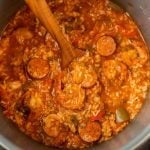A close-up of an instant pot shrimp jambalaya, featuring shrimp, sliced sausage, rice, diced tomatoes, and green peppers in a thick, spiced sauce, with a wooden spoon stirring the mixture.
