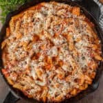 A cast iron skillet filled with baked pepperoni chicken pasta, tender chicken pieces, tomato sauce, and melted cheese, topped with herbs.