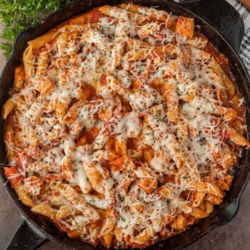 A cast iron skillet filled with baked pepperoni chicken pasta, tender chicken pieces, tomato sauce, and melted cheese, topped with herbs.